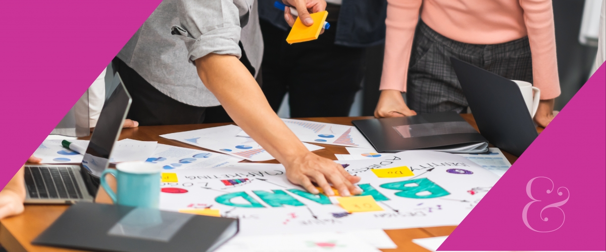 People are standing and working around a table. There are sticky-notes, computers, books and posters. A poster says "brand."