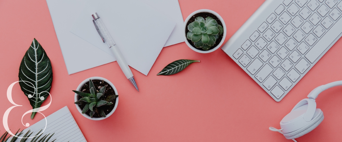 Dark green plants, office supplies, and a keyboard and headphones sit on top of a pink desk. A white ampersand is in the bottom left corner. 