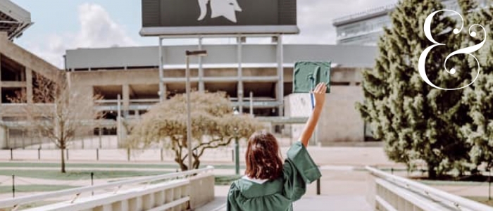 Image features a student in a green cap and gown in front of the MSU football stadium. 